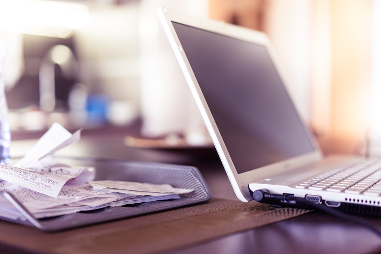 Close-up of an open laptop beside a mesh tray filled with receipts on a desk