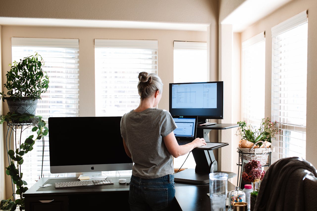 Woman in a gray shirt working at a standing desk with dual monitors in a bright home office