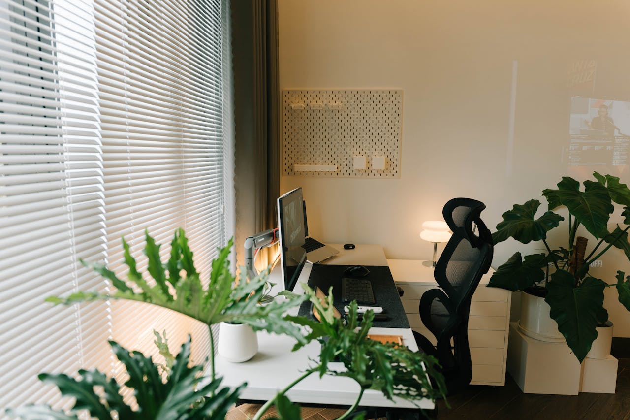 Dedicated home office workspace with desk, monitor, and plants beside a window with blinds